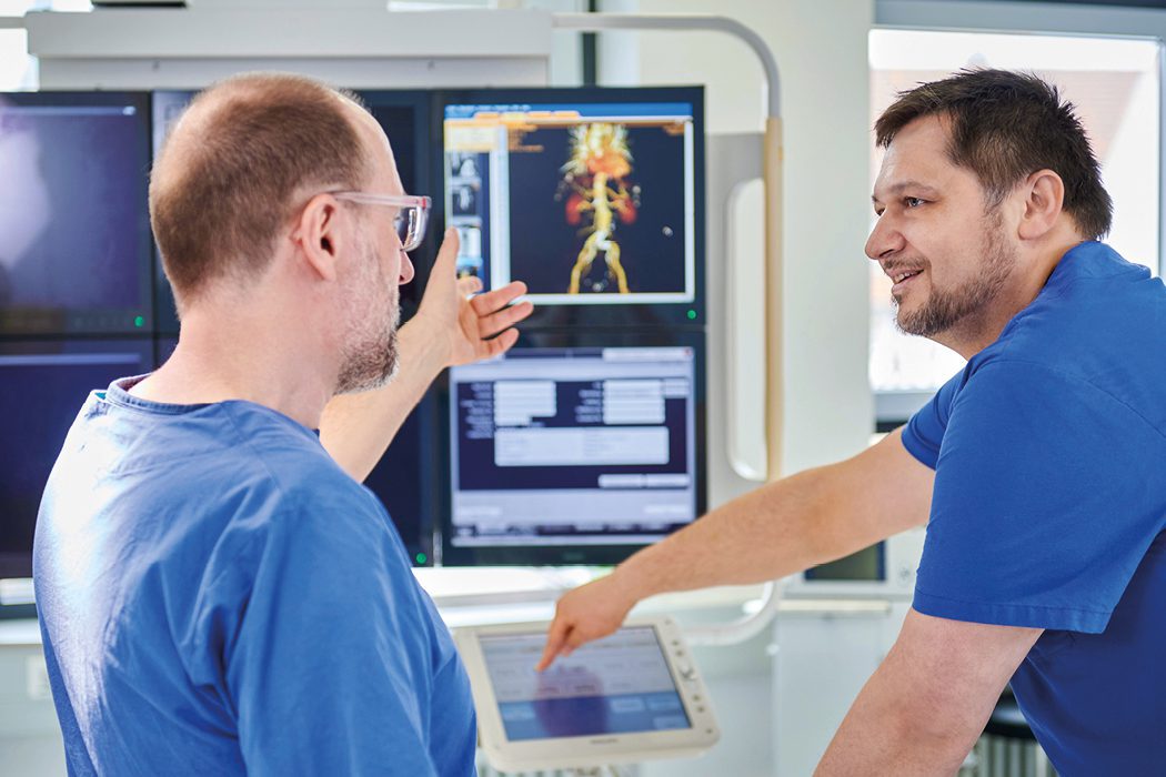 Gefäßchirurgen in blauer Arbeitskleidung besprechen sich vor einem Monitor.