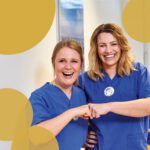 zwei lachende weibliche Pflegekräfte in blauer Krankenhausarbeitskleidung mit gelben Kreisen