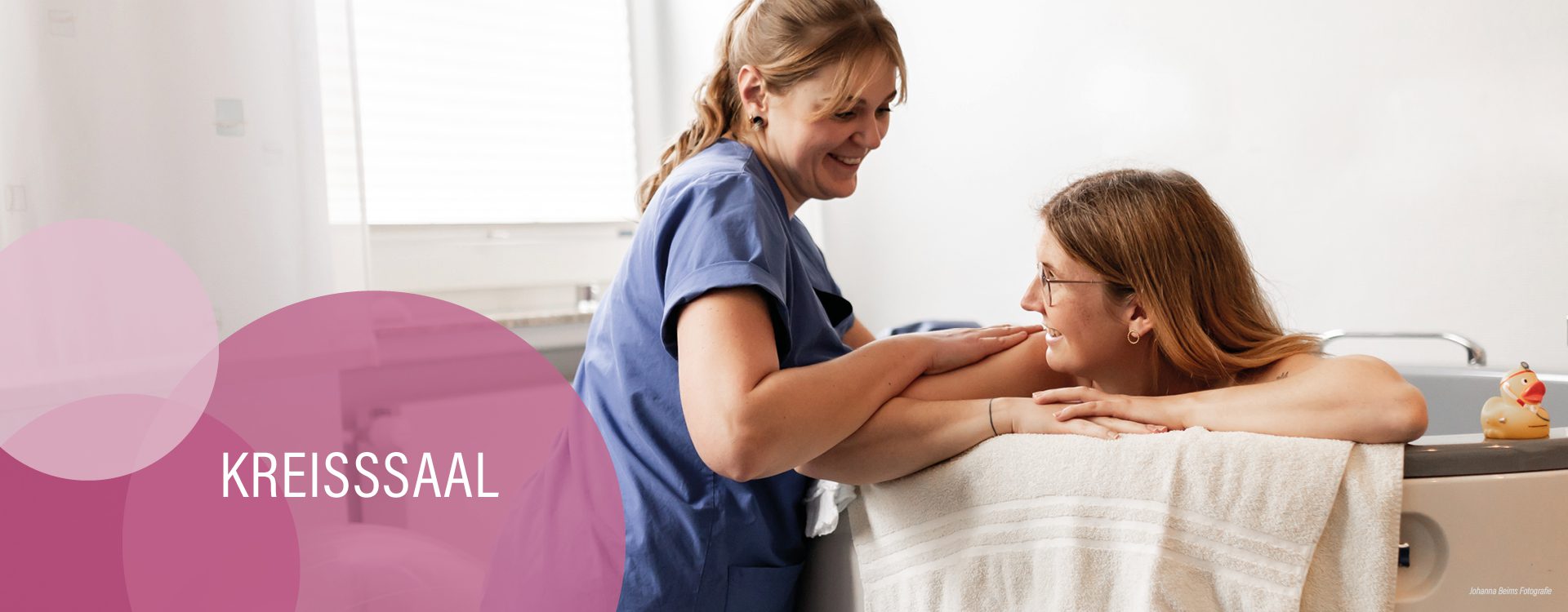 Eine Krankenschwester lächelt und stützt eine Frau in den Wehen, die sich auf den Rand einer Gebärwanne stützt, beide wirken glücklich und ruhig. Das Wort KREISSSAAL ist links auf einem rosa Overlay zu sehen.