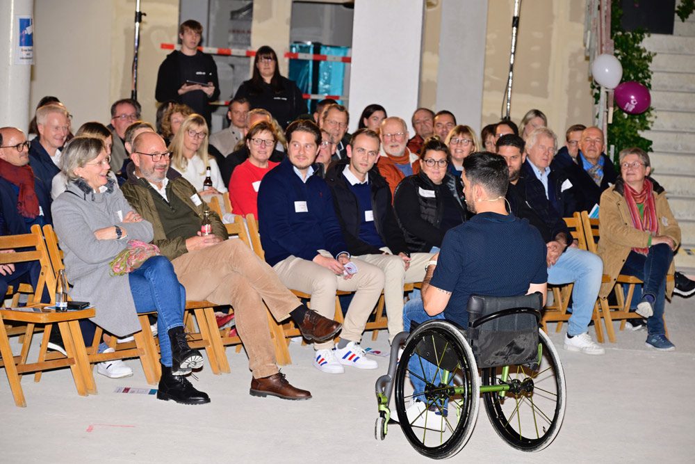 Ein Mann im Rollstuhl spricht zu einem aufmerksamen Publikum, das auf Holzstühlen in einem Innenraum sitzt. Die Zuhörer sind in einem hell erleuchteten Raum mit Luftballons und sichtbaren Treppen im Hintergrund beschäftigt, einige lächeln.