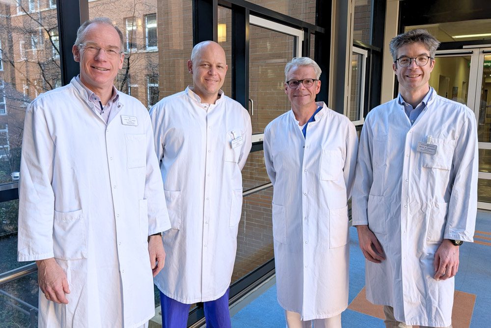 Vier Männer in weißen Laborkitteln stehen zusammen in einem hellen Flur mit großen Fenstern. Sie lächeln und scheinen medizinische Fachkräfte oder Wissenschaftler zu sein.