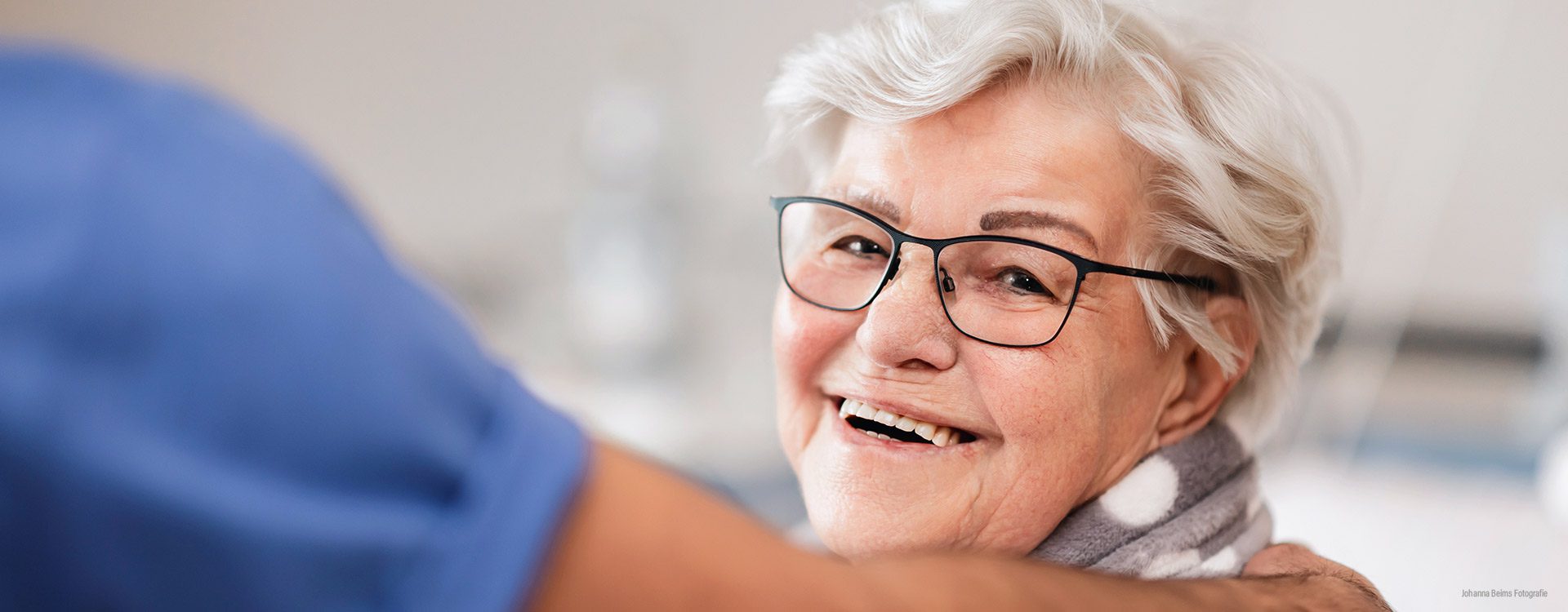 Eine ältere Frau mit kurzen weißen Haaren und einer Brille lächelt warmherzig. Eine Person in einem blauen Kittel berührt sie sanft an der Schulter, was auf eine fürsorgliche Interaktion oder Unterstützung hindeutet.
