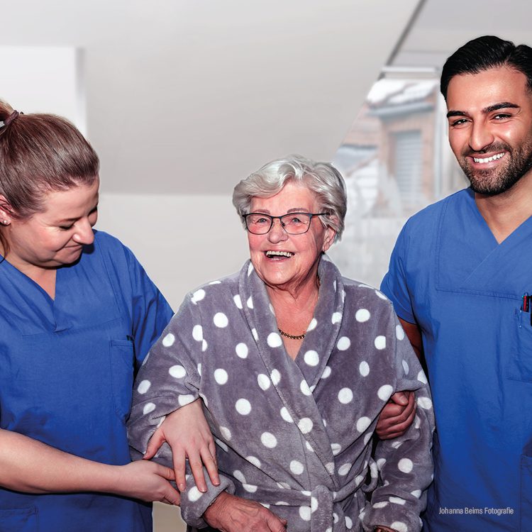 Zwei Pflegekräfte in bleuen Kasacks unterstützenm eine ältere Dame mit weißem Haar beim gehen.