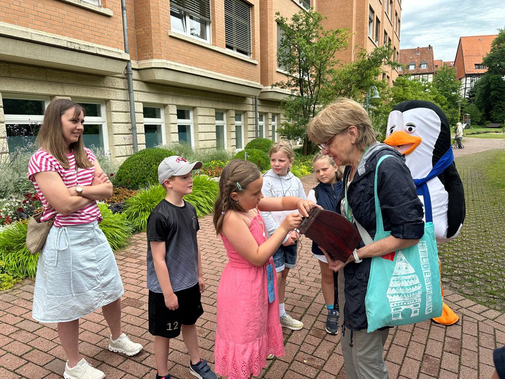 Eine Frau, die eine Tasche in der Hand hält, lächelt, während ein kleines Mädchen in einem rosa Kleid ihr etwas zeigt; daneben stehen drei Kinder und eine weitere Frau. Im Hintergrund steht eine Person in einem Pinguinkostüm. Sie befinden sich draußen in der Nähe eines Backsteingebäudes.