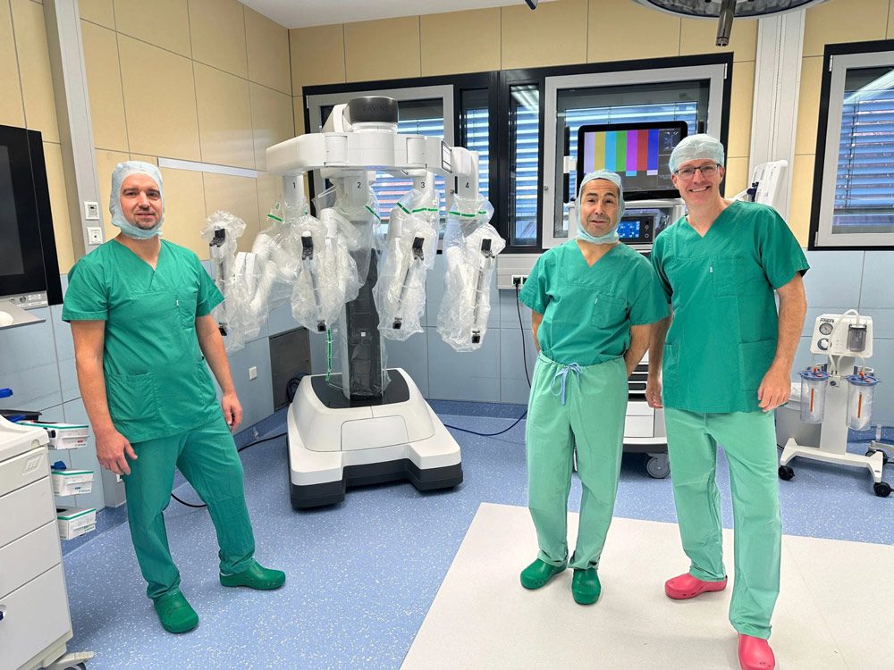 Drei Chirurgen in grüner OP-Kleidung stehen in einem Operationssaal neben einem chirurgischen Robotersystem. Im Hintergrund sind medizinische Geräte und Monitore zu sehen.
