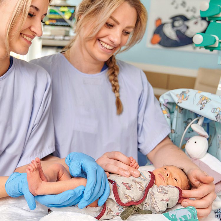 Zwei freundliche weibliche Pflegefachkräfte in hellblauer Arbeitskleidung versorgen ein Frühgeborenes in seinem Bettchen