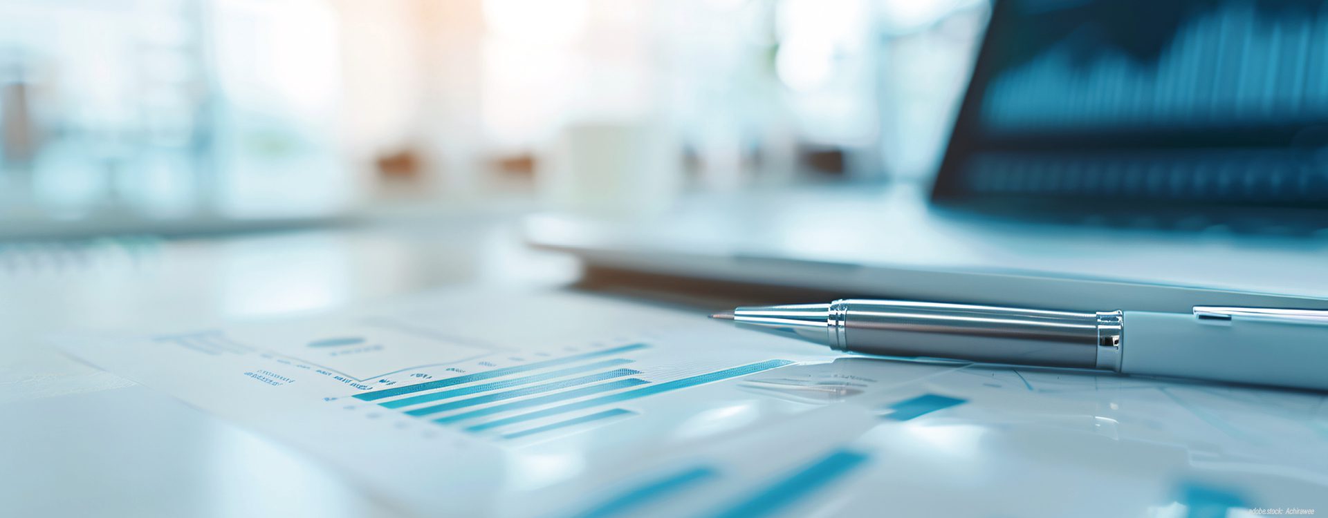 ein aufgeklappter Laptop auf einem Tisch mit Papieren und mit einem Schreibstift im Vordergrund