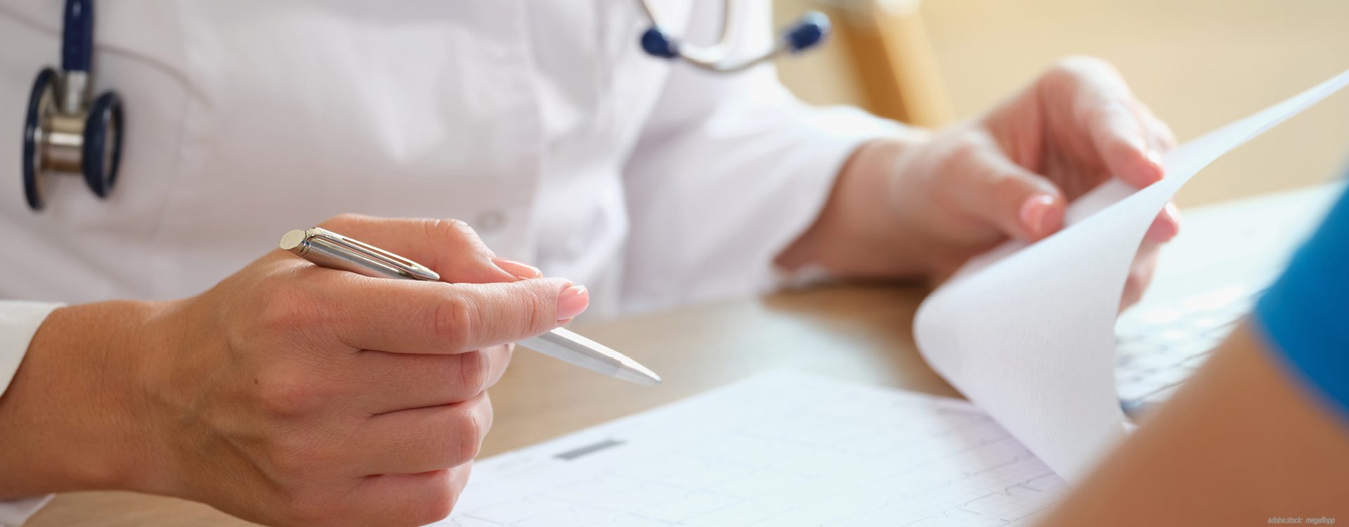 eine Person mit weißem Kittel sitzt an einem Tisch. Sie hält in ihrer rechten Hand einen Kugelschreiber und blättert mit der linken Hand ein Blatt Papier auf. Vor ihr sitzt eine weitere Person die aber nur zu erahnen ist.