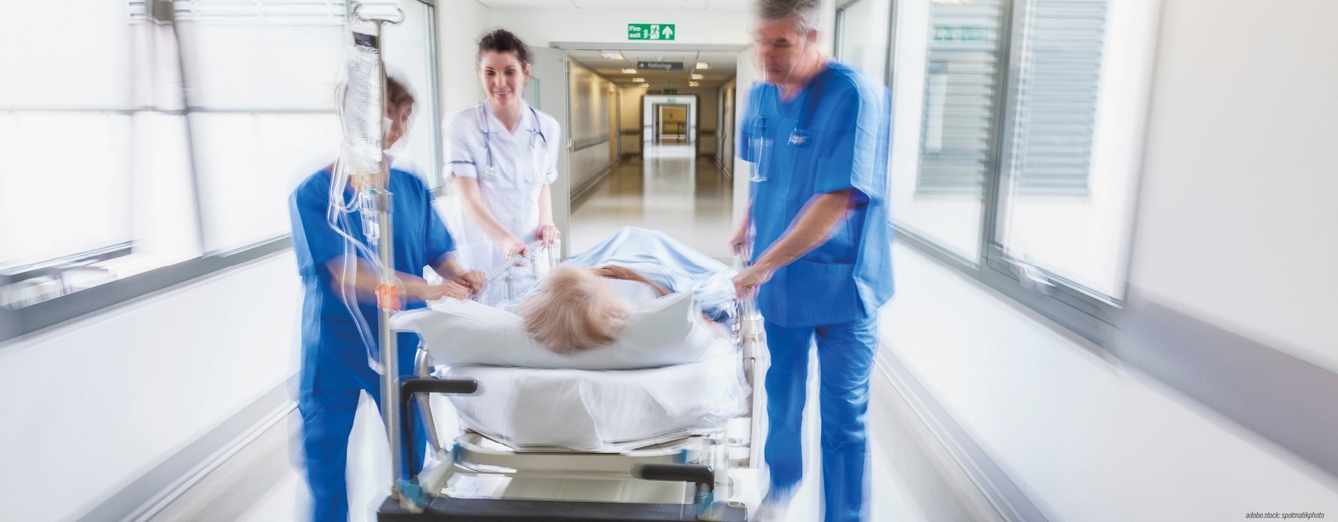 Drei Personen in Krankenhausarbeitskleidung schieben eine Trage mit einer liegenden Person einen Krankenhausflur entlang.