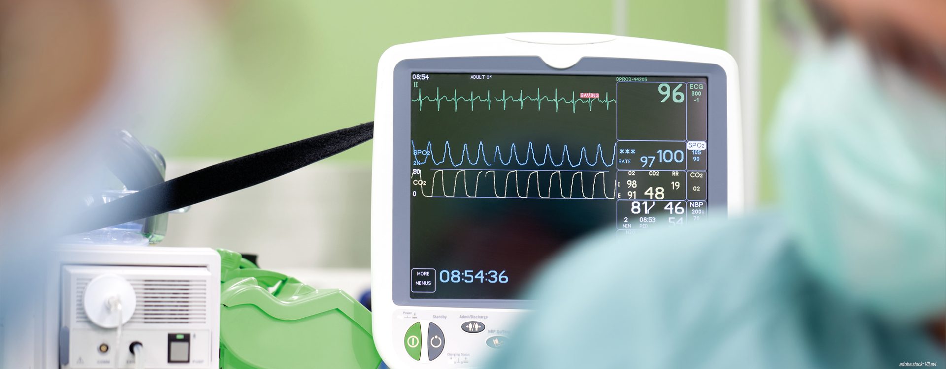 unscharf dargestellte Person in grüner Krankenhausarbeitskleidung vor einem Überwachungsmonitor