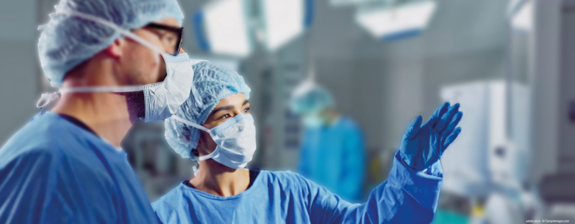 Zwei medizinische Fachkräfte in bleuer OP-Arbeitskleidung blicken auf ein nicht erkennbares Objekt an der Wand, im Hintergrundeine OP-Situation