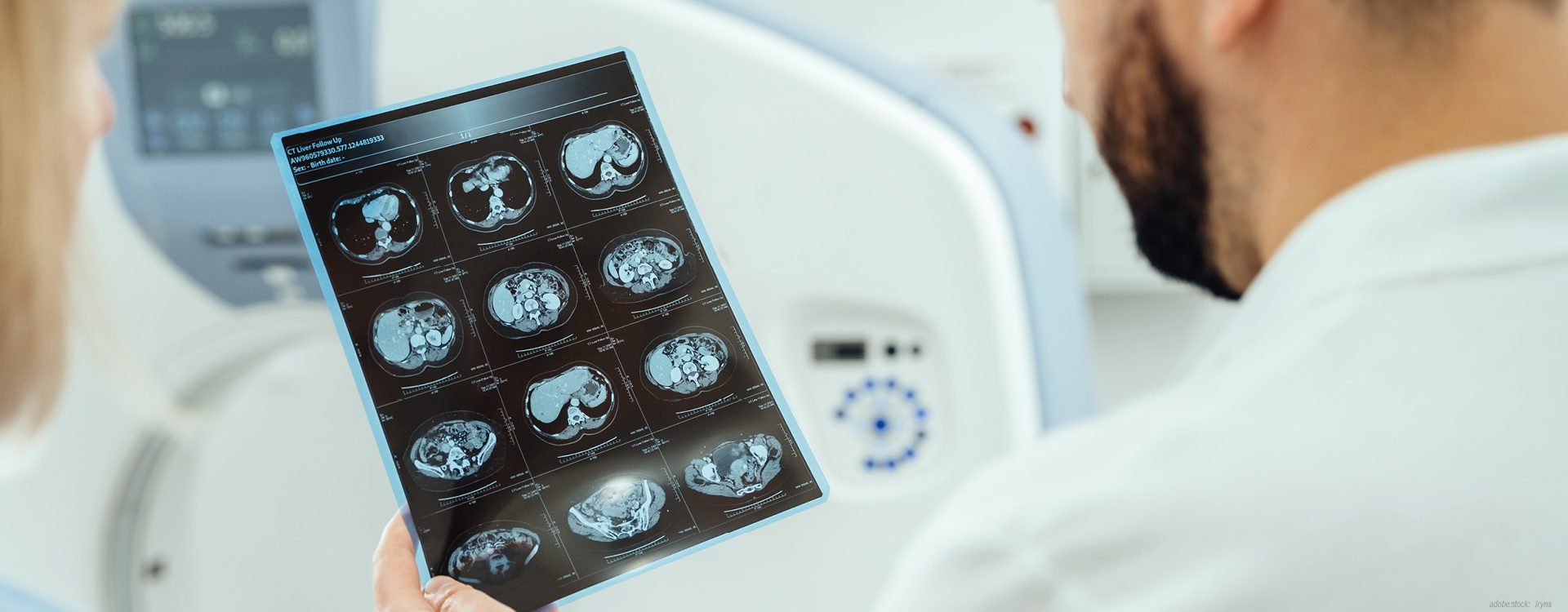 zwei Personen in weißen Kitteln stehen mit dem Rücken zum Betrachter und sehen ein CT-Bild mit mehreren Aufnahmen von einem menschlichen Gehirn an