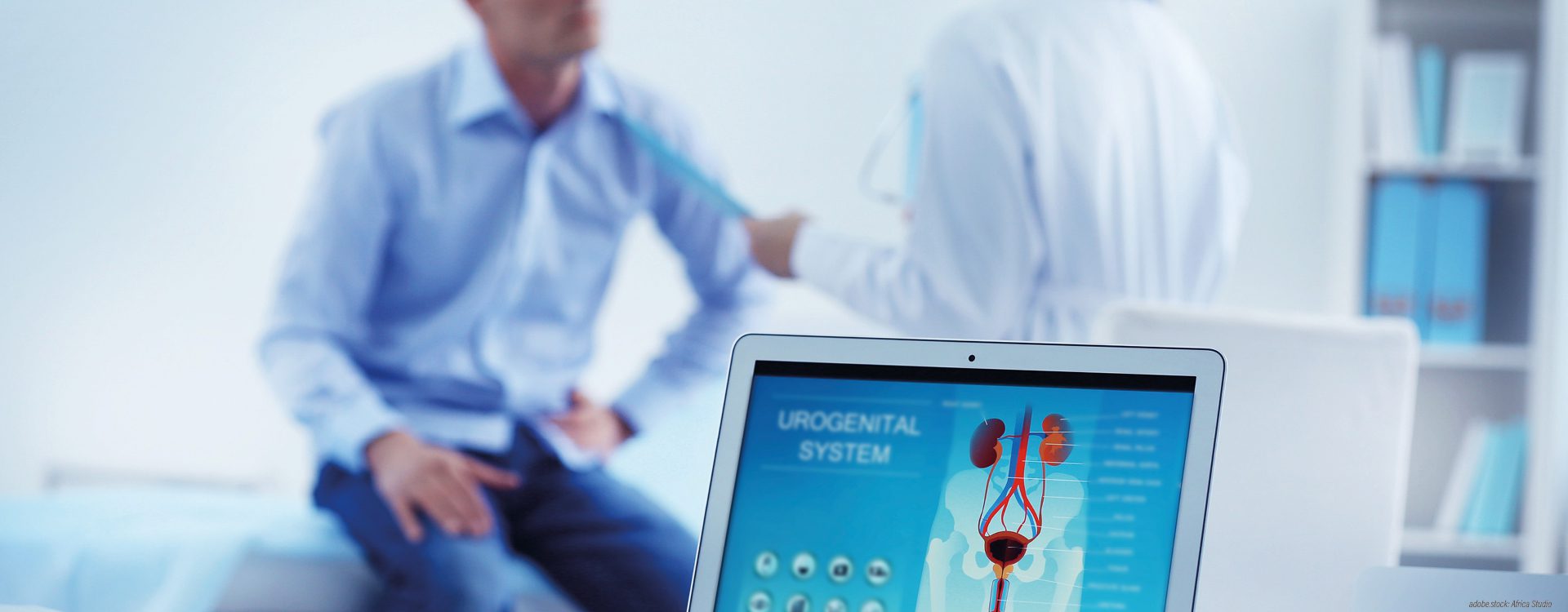 unscharf abgebildeter Mann sitzt auf einer Praxisliege, seine Ärztin oder Arzt erklärt ihm etwas, im Vordergrund ein aufgeklappter Laptop, der Bildschirm zeigt den männlichen Harntrakt