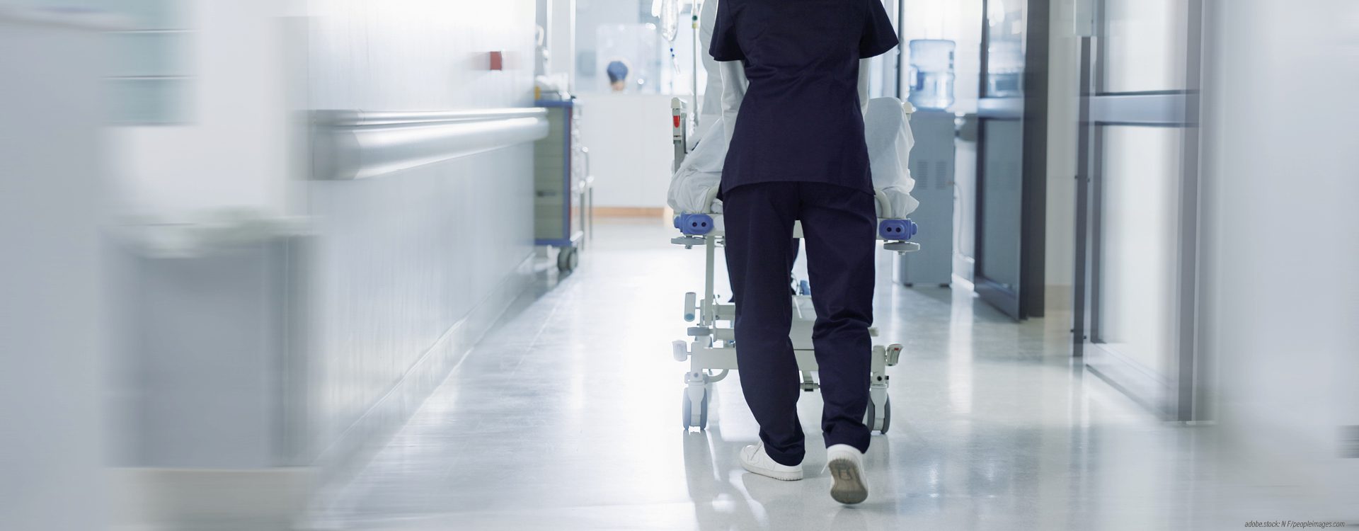Person in blauer Krankenhausarbeitskleidung schiebt eilig eine Trage mit einer liegenden Person einen Krankenhausflur entlang