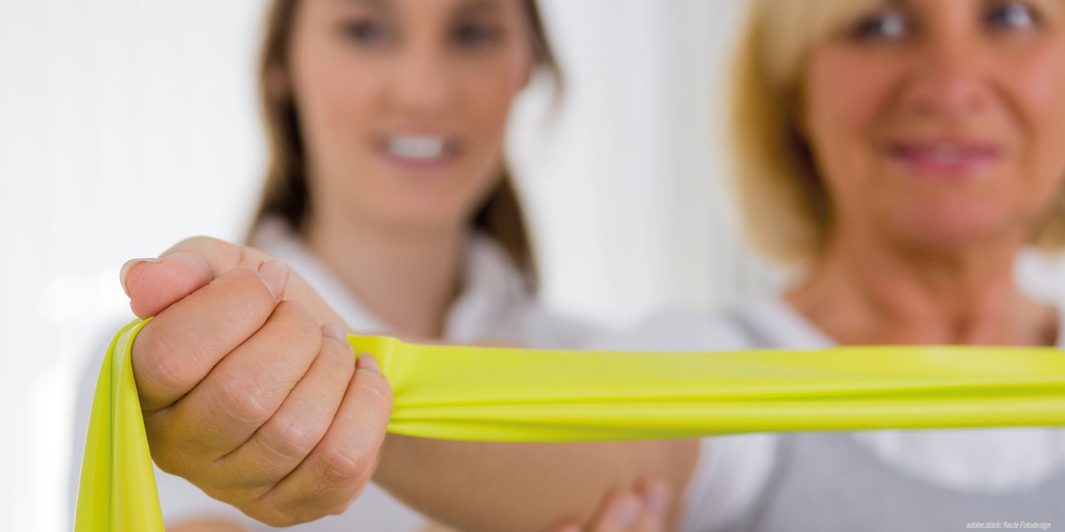 Zwei lächelnde, unscharf frontal abgebildete weibliche Personen. Im Vordergrund eine Patientin mit gelbem Stretchband, die Physiotherapeutin steht hinter ihr und unterstützt sie