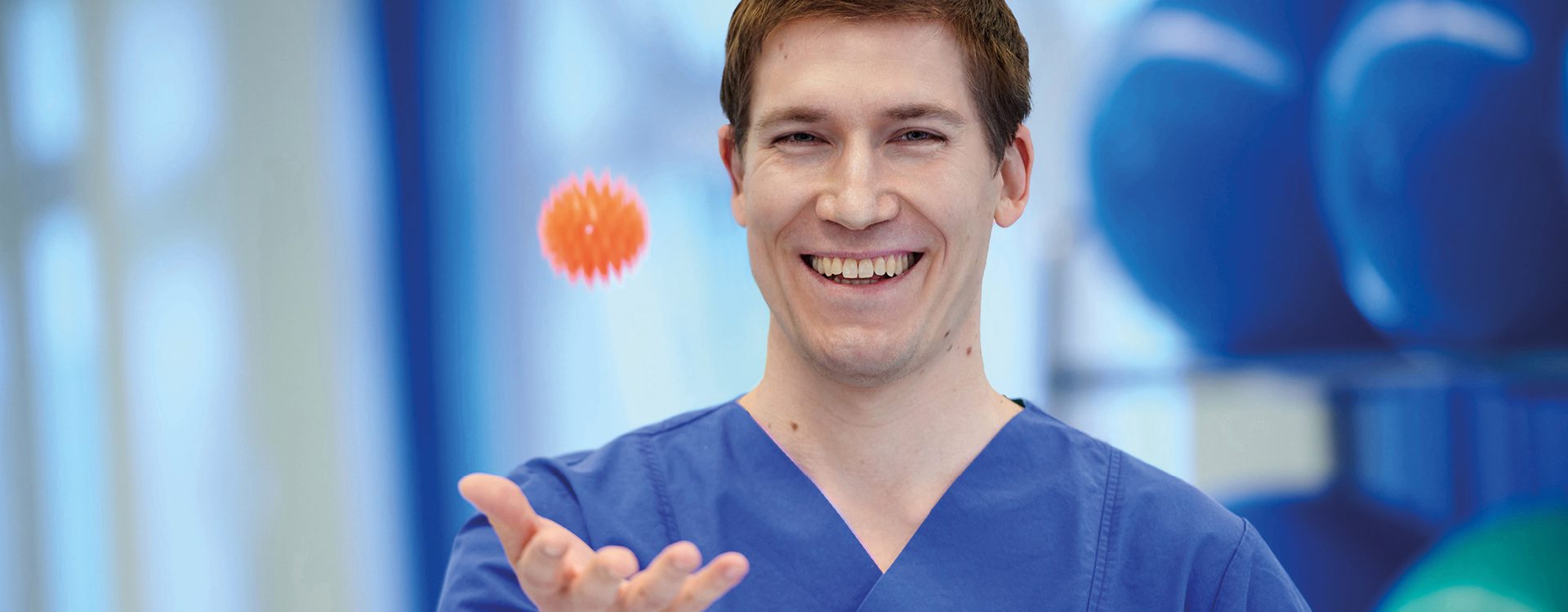 Ausschnitt einer freundlichen, männliche Person in blauer Krankenhauskleidung, fängt einen Stressball mit ausgestreckter Hand