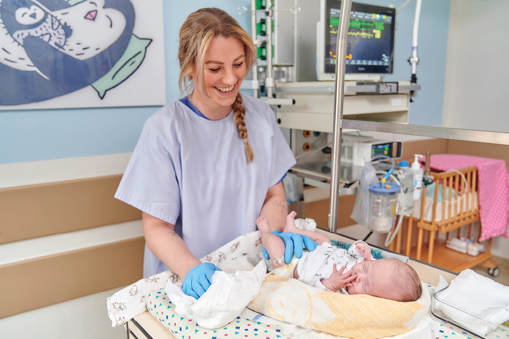 freundliche weibliche Pflegefachkrafte in hellblauer Arbeitskleidung versorgen ein Frühgeborenes in seinem Bettchen